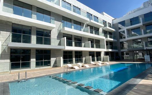 Un moderno complejo de apartamentos en Dubai con una gran piscina en el patio, rodeado de edificios blancos de varias plantas bajo un cielo azul despejado.