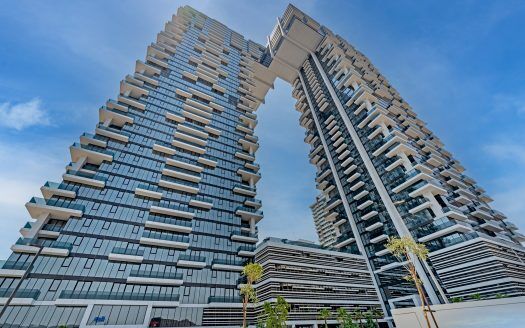 Edificios de arquitectura moderna con un singular puente elevado que une dos rascacielos en Dubai, bajo un cielo azul despejado. El diseño incluye varios balcones y una estructura en capas.
