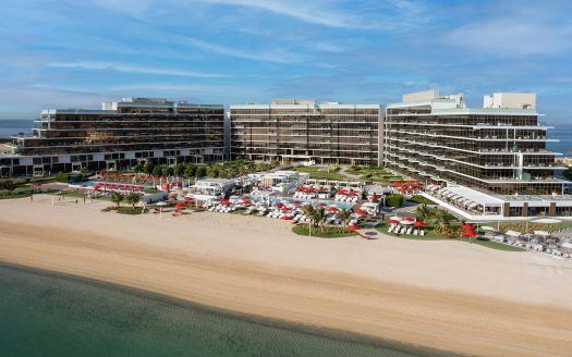 Vista aérea de un moderno complejo turístico de varias plantas en primera línea de playa con sombrillas rojas y tumbonas en una playa de arena adyacente de tranquilas aguas azules, ideal para invertir en Dubai.