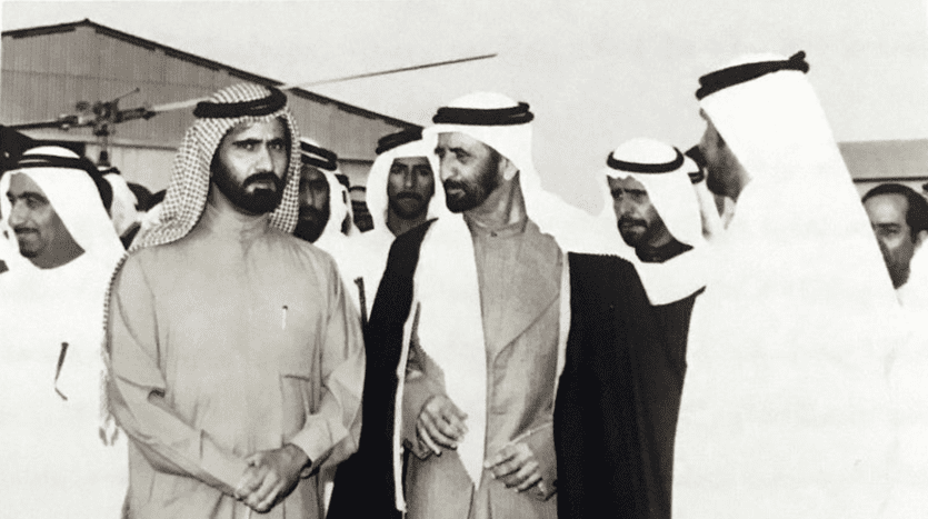 Photo en noir et blanc d'un groupe d'hommes du Moyen-Orient en tenue traditionnelle engagés dans une conversation sur l'immobilier Dubaï, debout à l'extérieur avec des bâtiments visibles en arrière-plan.