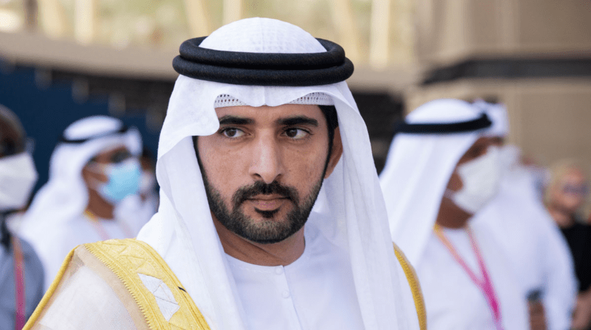 Un homme vêtu d'une tenue traditionnelle émiratie, comprenant une kandura blanche et une ghutra noire et blanche, regarde pensivement devant lui un événement en plein air près d'une villa à Dubaï.