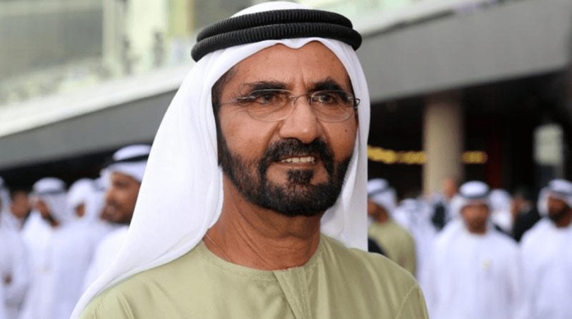 Un homme vêtu d'une tenue traditionnelle émiratie, comprenant un kandura blanc et un agal noir, se tient dans un quartier de villas bondé à Dubaï, souriant légèrement à la caméra.