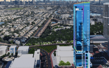 Vue aérienne d'un paysage urbain présentant un grand gratte-ciel moderne doté d'un éclairage LED coloré, entouré de bâtiments plus petits et de zones résidentielles. En arrière-plan, d’autres immeubles de grande hauteur et un front de mer sont visibles, avec un ciel clair et expansif au-dessus.