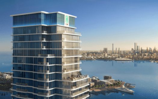 Un moderno rascacielos con ventanas de cristal, el LIV Maritime, ocupa un lugar destacado en el paseo marítimo. El horizonte de la ciudad y un famoso rascacielos se ven al fondo en un cielo azul despejado.