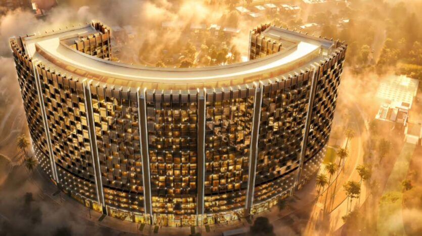 Une vue aérienne de l'impressionnant Binghatti Wraith, un immense bâtiment moderne en forme de U à la façade dorée, entouré d'arbres et de brume, tandis que les rayons du soleil y pénètrent pour créer une atmosphère chaleureuse et lumineuse.