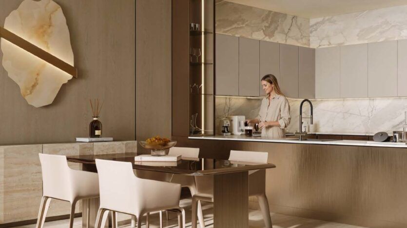 Une femme prépare du café dans une cuisine moderne Binghatti Wraith aux tons beiges. La pièce se caractérise par des murs en marbre, des meubles épurés, une table à manger avec quatre chaises et un éclairage doux et chaleureux. Un panneau mural décoratif et une corbeille à fruits apportent une touche d'élégance.
