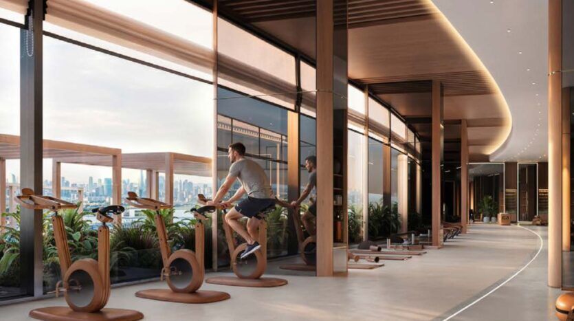 Une salle de sport moderne au Binghatti Wraith, dotée de grandes baies vitrées donnant sur les toits de la ville, où l'on voit un homme sur un vélo d'appartement. L'espace est lumineux, avec des touches de bois, des plantes vertes et de nombreux appareils de musculation.
