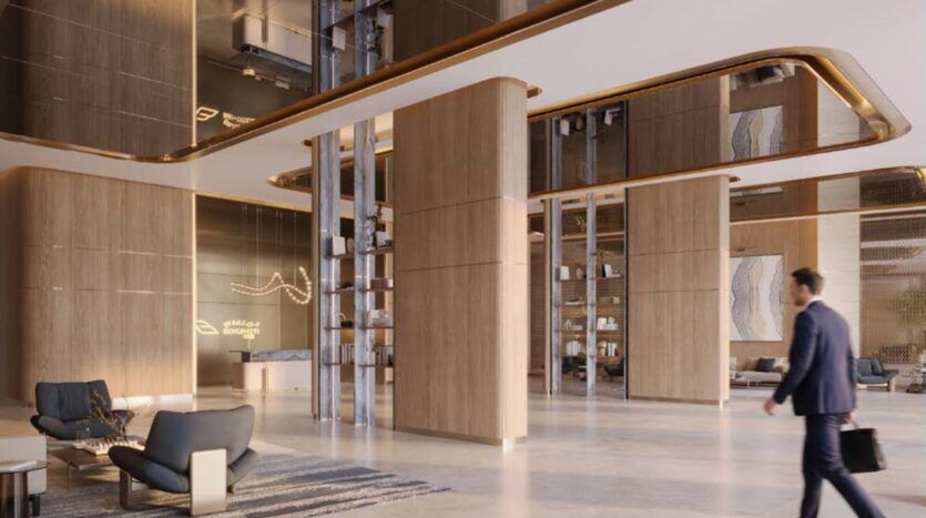 Un hall d'entrée moderne aux hauts plafonds, rehaussé de touches de bois et de métal, et doté d'un coin salon élégant. Un homme en costume, un sac à la main, traverse cet espace inspiré de la Binghatti Wraith, où un éclairage d'ambiance chaleureux crée une atmosphère accueillante.