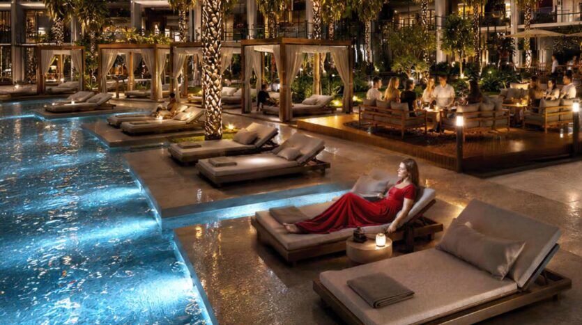 Une femme vêtue d'une robe rouge se détend sur une chaise longue au bord d'une piscine illuminée de nuit, entourée de palmiers éclairés, de cabanes et de convives dînant en plein air dans l'ambiance somptueuse du complexe Binghatti Wraith.