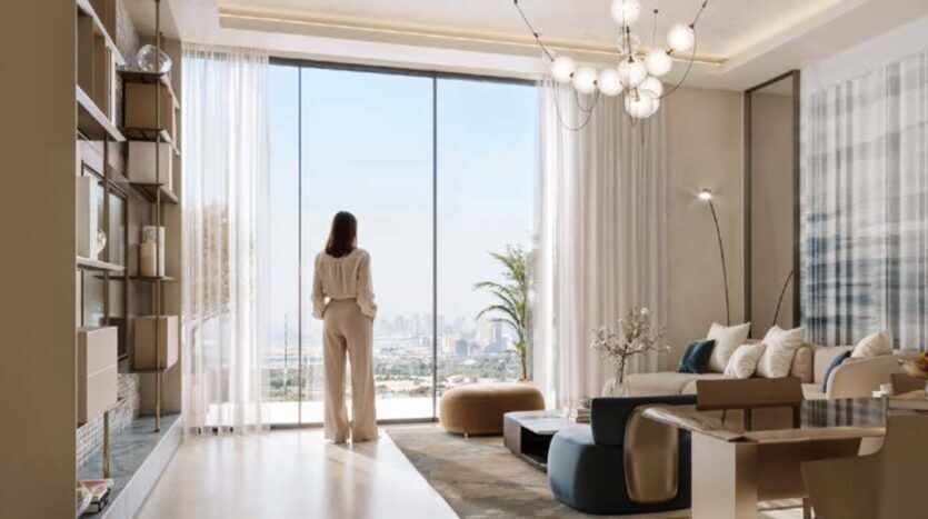 Une femme se tient près des baies vitrées d'un salon moderne conçu par Binghatti Wraith, contemplant le paysage urbain. L'espace se caractérise par un mobilier contemporain, des tons neutres et une abondance de lumière naturelle.