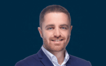 Un portrait professionnel d'un homme souriant avec une barbe, vêtu d'un blazer bleu marine et d'une chemise blanche, sur un fond bleu sur le thème "immobilier Dubaï".