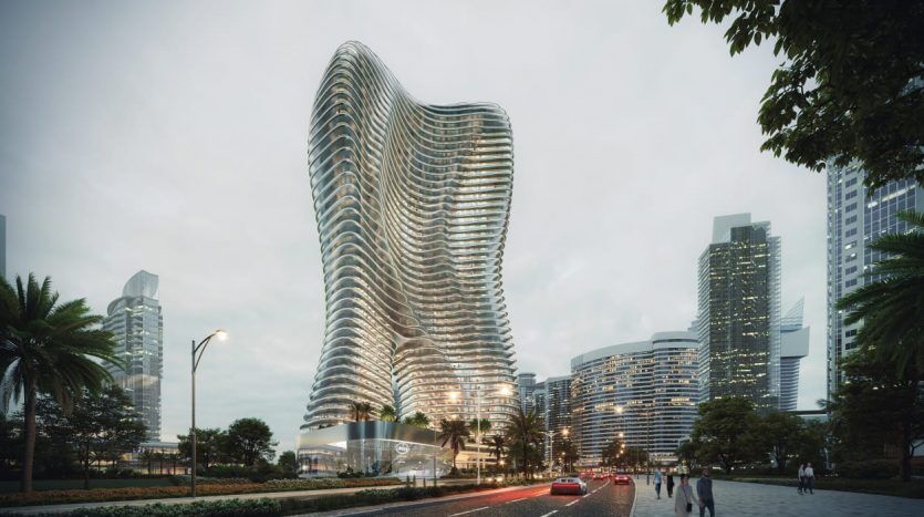 Image architecturale d'un gratte-ciel moderne et ondulé avec des surfaces en verre réfléchissantes à Dubaï, entouré d'arbres et éclairé le soir. Des voitures passent sur la route voisine sous un ciel nuageux.