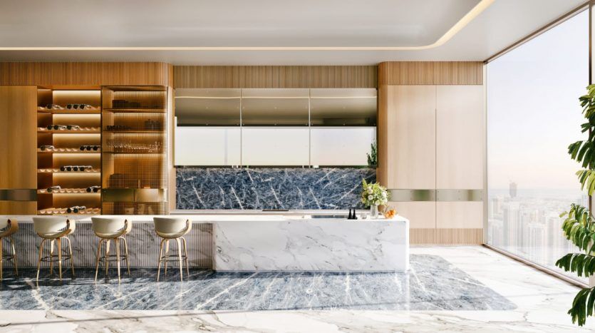 Une cuisine moderne et luxueuse dans un appartement de Dubaï avec des comptoirs et un sol en marbre, des armoires en bois et une vue sur la ville à travers de grandes fenêtres. Il y a un coin salon de style bar avec plusieurs tabourets.