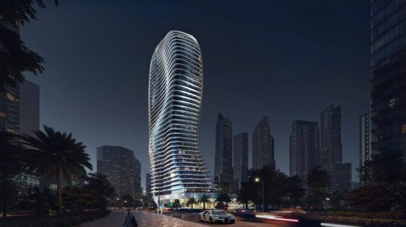 Un gratte-ciel futuriste de Dubaï, illuminé sous un ciel nocturne, se détache sur fond de grands immeubles et d'arbres luxuriants. Une voiture floue passe, créant une traînée lumineuse dans la rue.