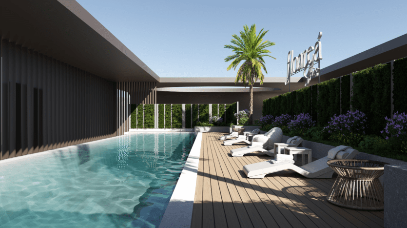 Une piscine extérieure moderne aux eaux claires, entourée de chaises longues, de tables d'appoint et de plantes en pot. Un palmier et des arbustes à fleurs bordent la terrasse sous un ciel fleuri, avec un panneau « Aquéa » en arrière-plan.
