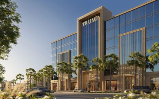 Un bâtiment moderne à la façade vitrée, avec des palmiers à l'avant et la mention "TRUMP PLAZA" au sommet. Des voitures passent dans la rue et le ciel est clair et bleu.