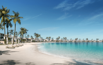 Une station balnéaire tranquille de Dubaï avec des palmiers, des villas modernes bordant le rivage et des eaux bleues claires sous un ciel lumineux. Des chaises longues sont disséminées le long de la plage de sable.