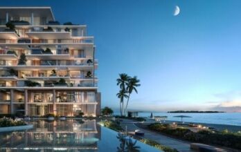 A modern beachfront building in Silena Dubai Islands features glass balconies overlooking a calm pool, palm trees and a sandy beach at dusk, under a crescent moon. The sky changes from deep blue to pale along the horizon.