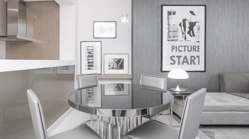 Modern dining area in Damac Towers with glass table, four chairs and framed artwork on gray walls. The space opens onto a kitchen and features minimalist decor in muted tones.