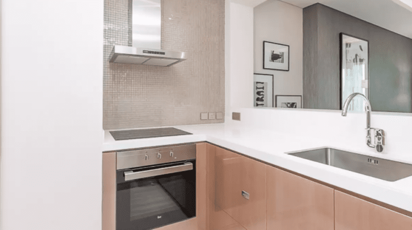 Modern kitchen in Damac Towers Paramount with wood cabinets, stainless steel built-in oven, white countertops and mosaic backsplash. An elegant hood sits above the stove.