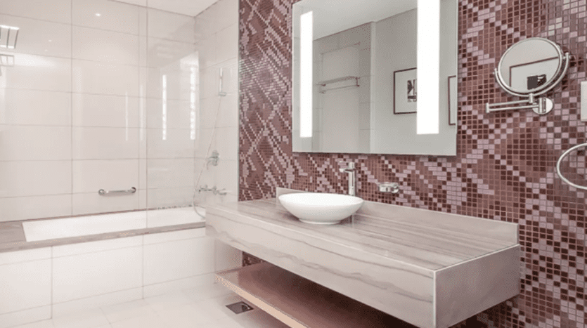Modern bathroom in Damac Towers with red mosaic tiles, floating wooden vanity with white sink and large mirror above. The floor is tiled and there's a glass shower cubicle in the