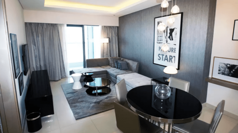 The living room of a modern apartment at Damac Towers Paramount features a gray sofa, glass dining table with chairs, framed artwork on the wall and ambient lighting.