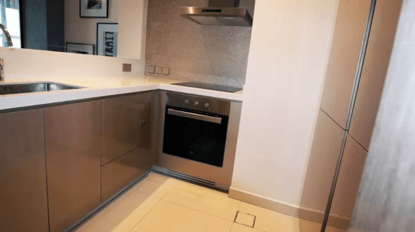 Contemporary kitchen area in Damac Towers Paramount featuring elegant brown cabinetry, built-in oven and stainless steel hood, with mosaic backsplash and tiled floor.