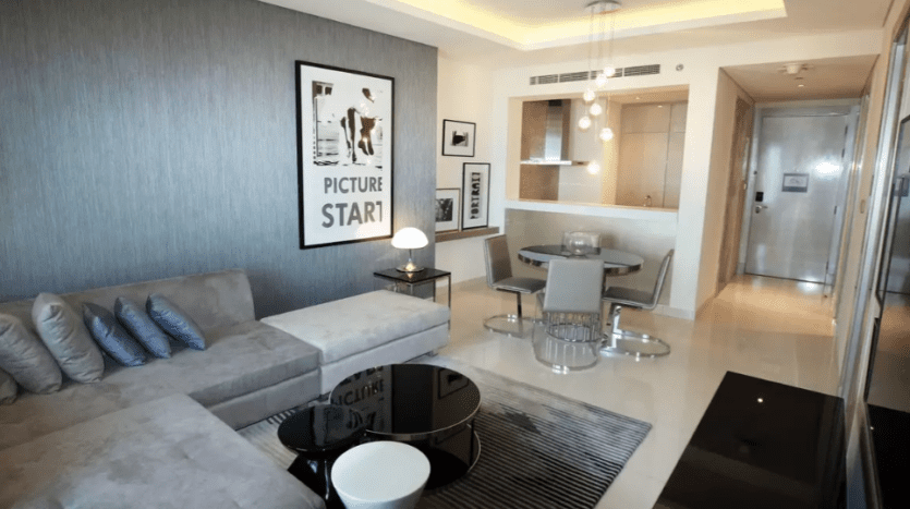A modern living room in Damac Towers with a gray sectional sofa, black coffee table, dining area in the background and elegant decor with framed wall art.