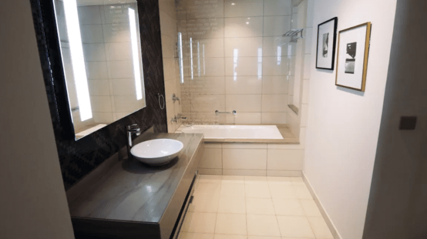 Modern bathroom interior in Damac Towers featuring a large mirror, white bathtub with glass enclosure, wall-mounted sink and framed photographs on the wall. Bright, uncluttered design.