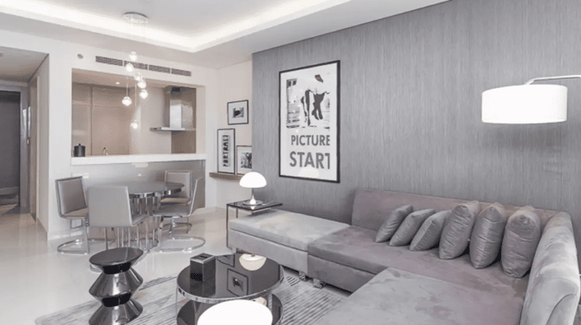 Modern living room with gray sofa, black side tables, white lamp and dining area in the background at Damac Towers Paramount. The walls feature minimalist decor and an "image beginning