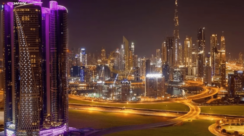 Night view of a well-lit cityscape with modern skyscrapers, including a tower with purple lighting, and curving, illuminated roads. The Burj Khalifa and Damac Towers Paramount are visible.