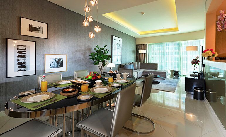 Modern dining area with an elegant glass table set for a meal, adjacent to a stylish living room and kitchen in Damac Towers, featuring contemporary furnishings and vibrant lighting.
