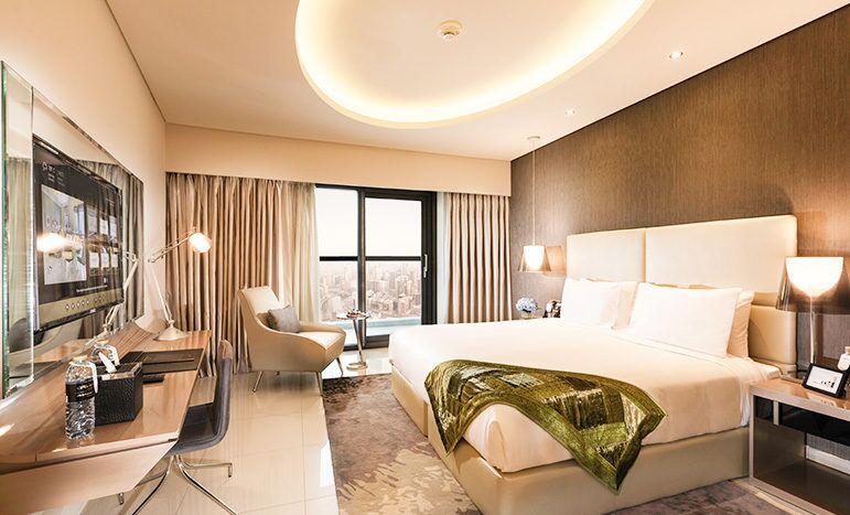 A modern hotel room in the Damac Towers featuring a king-size bed with a green throw, a round ceiling light, a desk area, an armchair and a view of the city through the window. Neutral tones and contemporary design enhance the room.
