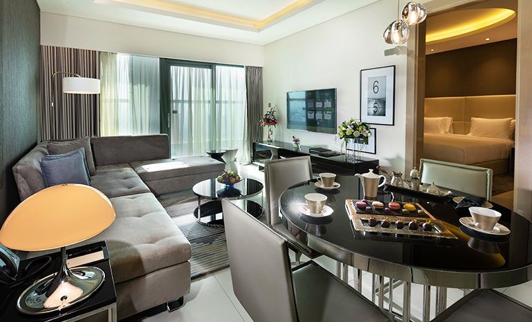 Modern hotel room interior at Damac Towers Paramount featuring a stylish lounge area with gray sofa, glass coffee table and adjoining dining area, all under warm ambient lighting.