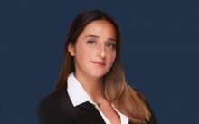 Portrait professionnel d'une femme aux longs cheveux bruns portant un blazer noir sur une chemise blanche, idéal pour une agence immobilière de Dubaï, sur fond bleu.