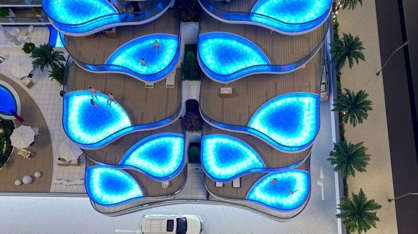 Vue aérienne du toit d'un bâtiment présentant un arrangement de piscines éclairées en bleu ressemblant à des pétales de fleurs, entourées de palmiers verts à Samana Park Views et d'une zone pavée blanche avec une voiture garée.