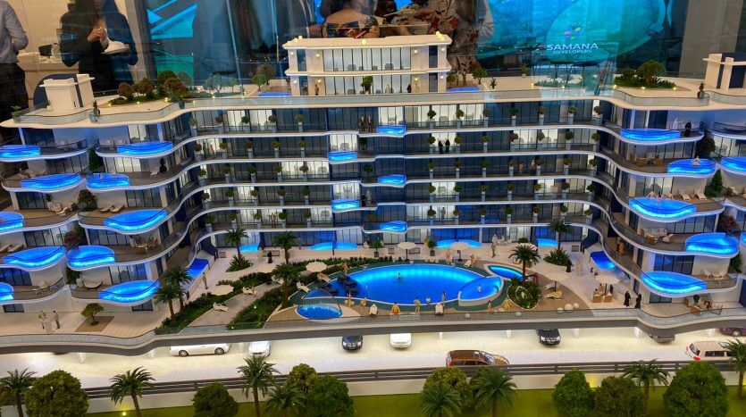 Un modèle détaillé de Samana Park Views, un complexe d'appartements moderne avec une piscine centrale, des balcons éclairés en bleu, des palmiers environnants, des voitures miniatures sur la route et des personnes examinant le modèle