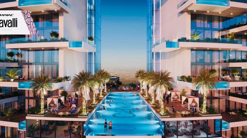 A luxurious pool between two high-rise buildings, lined with palm trees and cabanas, under a clear sky. People relax and swim in the water. Logos for Damac Tower and Roberto Cavalli