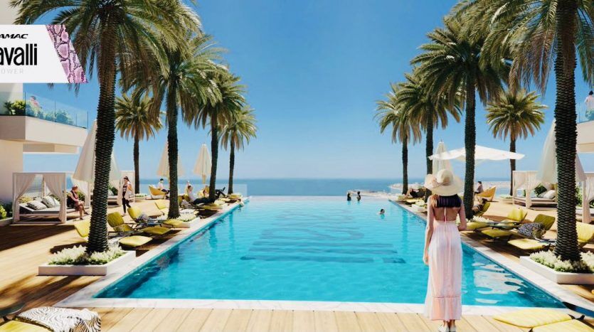 A woman wearing a hat and flowing dress stands at the edge of a luxurious palm-fringed pool in Dubai's Roberto Cavalli Tower, overlooking the sea. Sun loungers and parasols dot the pool deck.