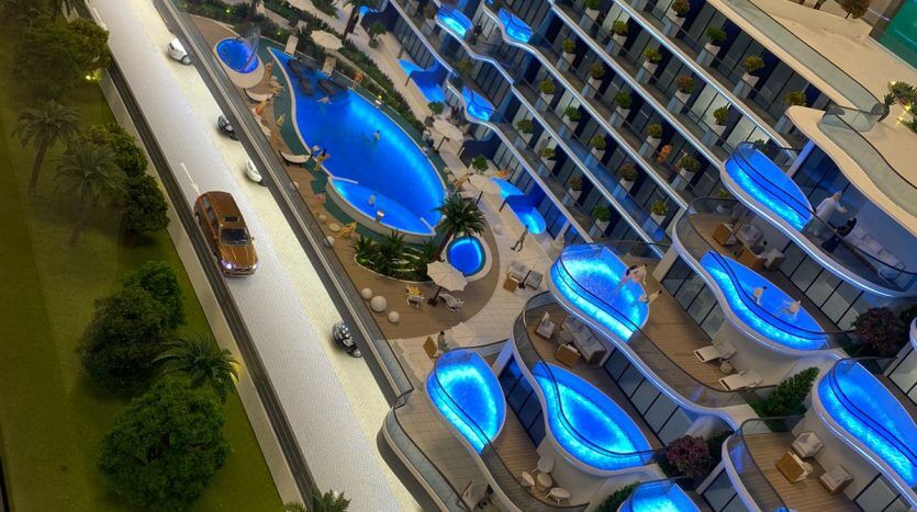 Une maquette du complexe résidentiel moderne Samana Park Views la nuit, avec des piscines illuminées en bleu, des jardins paysagers et des bâtiments environnants.