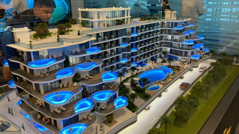 Une maquette détaillée d'un immeuble résidentiel moderne à plusieurs étages avec des balcons bleus éclairés, des terrasses paysagées et des piscines, exposée dans une salle d'une agence immobilière de Dubaï avec des spectateurs.