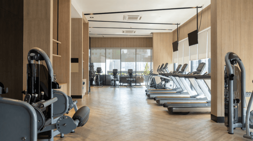 Intérieur d'une salle de sport moderne avec des équipements d'exercice comprenant des tapis roulants et des appareils de musculation, du parquet, de grandes fenêtres et une vue sur la ville de Binghatti Dubaï.