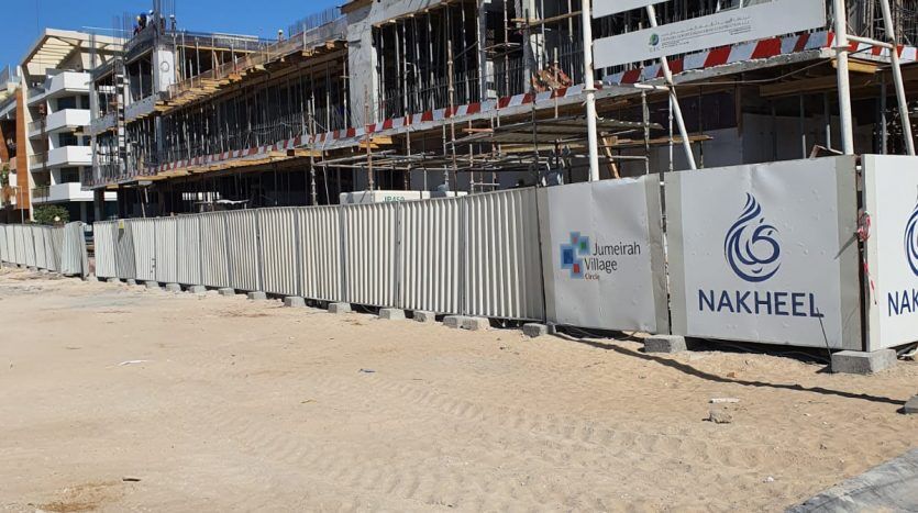 Un chantier de construction avec un grand bâtiment en cours de développement par Binghatti Dubaï, entouré d'une clôture métallique et comportant diverses signalisations d'entreprise. Un ciel bleu clair et un premier plan sablonneux sont visibles.