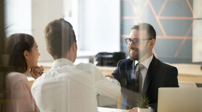 Trois professionnels, une femme et deux hommes, engagent une conversation sur le financement bancaire dans un bureau moderne avec des ordinateurs portables visibles sur la table.