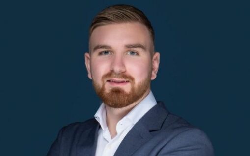 Une photo professionnelle d'un homme souriant avec une barbe, vêtu d'une chemise blanche et d'une veste de costume bleu marine sur un fond bleu sarcelle foncé, représentant une agence immobilière réputée de Dubaï.