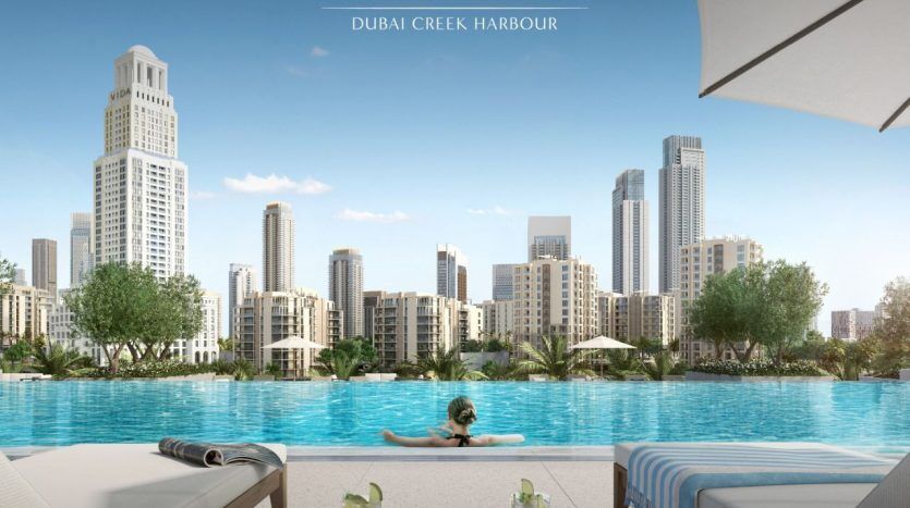 A woman swims in an infinity pool offering a panoramic view of Dubai Creek harbor, with modern high-rise apartment buildings under a clear blue sky.