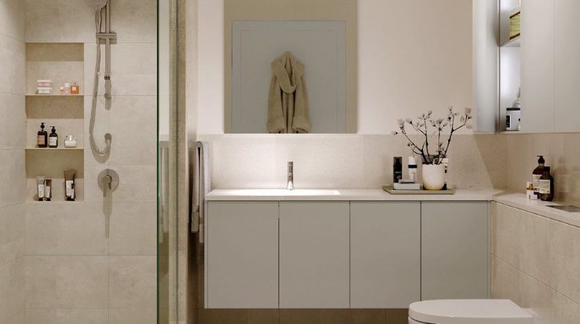 A modern bathroom with beige tiles and large glass shower. There's a white vanity with mirror, sink and decorative plants. A toilet rests next to a blue carpet. Text on image