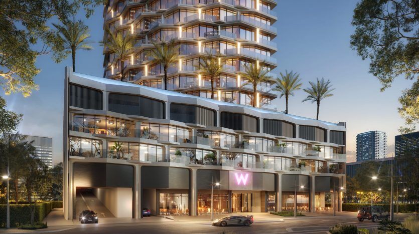 Vue extérieure d'un bâtiment moderne de plusieurs étages à Dubaï au crépuscule. Les lumières intérieures lumineuses révèlent un design luxueux avec des balcons en verre et un logo « w » au néon sur la façade. Des palmiers et un