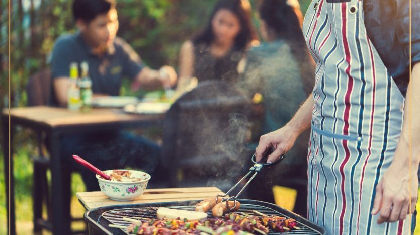 Un homme en tablier rayé fait griller un assortiment de viandes et de légumes sur un barbecue dans une villa de Dubaï, tandis qu'en arrière-plan, un groupe de personnes savoure un repas ensemble en plein air.