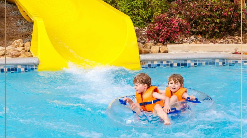 Deux enfants portant des gilets de sauvetage glissent sur un toboggan jaune sur un tube gonflable bleu et plongent dans une piscine entourée de rochers et de plantes à Dubaï.